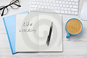 Phrase Life Coaching written in notebook, pen, cup of coffee, glasses and keyboard on white wooden table, flat lay