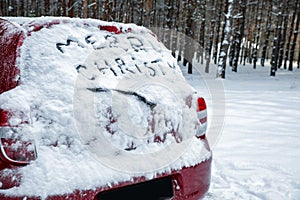 Phrase `Merry Christmas` written on snow covered car in winter forest.