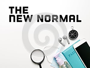 Phrase The New Normal on top of white desk with magnifying glass,sanitizer bottle,mask,passport,smartphone,ear buds,passport  and