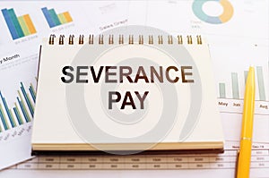 The phrase Severance Pay is written in a notepad that lies on a table with financial charts and diagrams. Business concept