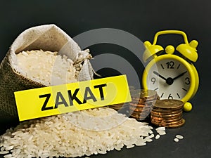 Phrase ZAKAT written on sticky note with rice in sack,coins and alarm clock.