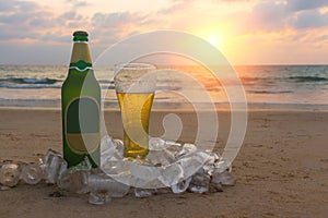 Misted bottle and glass of cold beer on the sand of scenic at the background of seascape, sunset sky and sea waves.