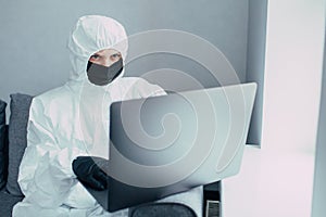 A physician in viral protective suit and face mask works on a laptop, online searching information about  coronavirus pandemic. Qu