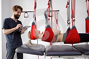 Physiotherapist doctor with clipboard watching female patient hanging on suspension system for neuromuscular activation.