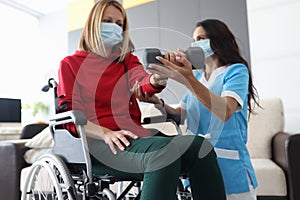 Physiotherapist doctor in medical mask helps to raise dumbbell to disabled woman in wheelchair at home portrait