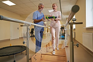 Physiotherapist helping mature patient walk between parallel bars in hospital