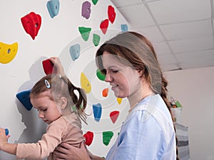 physiotherapist instructor helping little girl to climb wall in gym. sensory integration for kid and correctional