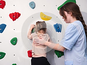 physiotherapist instructor helping little girl to climb wall in gym. sensory integration for kid and correctional