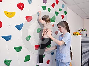 physiotherapist instructor helping little girl to climb wall in gym. sensory integration for kid and correctional