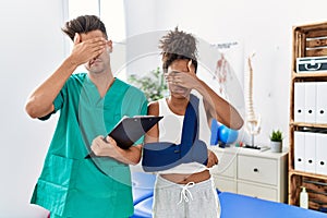 Physiotherapist working with patient wearing arm on sling at rehabilitation clinic covering eyes with hand, looking serious and