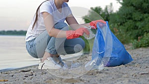 Picking garbage, volunteer woman in rubber gloves collects plastic trash in refuse bag while cleaning coast close up