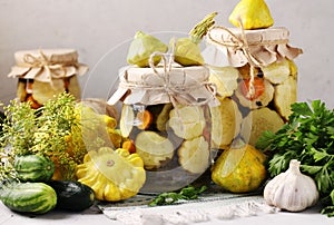 Pickled patissons, cucumbers with carrots and garlic in glass jars and fresh ingredients on a gray background