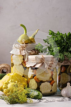 Pickled patissons, cucumbers with carrots and garlic in glass jars and fresh ingredients on a light gray background, Vertical