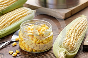 Pickled sweet corn in a glass jar, fresh corncobs, saucepan on table