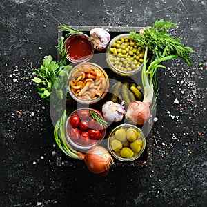 Pickled vegetables and mushrooms in glass jars in Wooden box on black stone background.