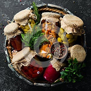 Pickled vegetables and mushrooms in glass jars in Wooden box on black stone background.