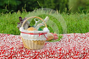 Picnic basket with a bottle of white wine, corkscrew, buns and bunch of basil on red tablecloth, plate with salad, tomat