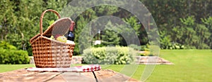 Picnic basket with fruits, bottle of wine and checkered blanket on table in garden, space for text. Banner design