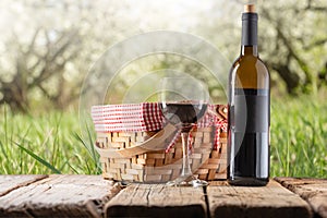 Picnic basket with red wine bottle and wine glass stands on wooden rustic table on green garden background. Concept of romantic