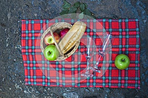 Picnic basket with wine glass and fruits on checkered cloth on rocks. Romantic still life of autumn or summer outing, lunch at