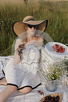 Picnic in nature on summer. Blonde in white dress, elegant hat drinks champagne with strawberries.