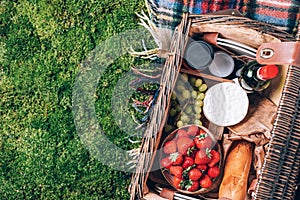 Picnic set with fruit, cheese, honey, strawberries, grapes, baguette, wine, wicker basket for picnic on plaid over green