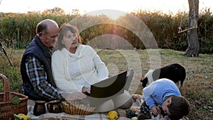 Picnic on sunrise, attractive mature couple with grandson and pet sits at plaid with laptop