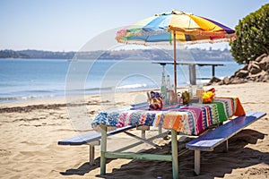 picnic table on the beach, with colorful umbrellas and towels, and sparkling water in the background