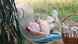 Picnic time. Happy young girl with round hat laying near the tree and using her mobile phone. Woman web surfing in the