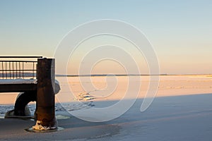 Pictorial winter landscape of frozen river lit by red sun and endless cloudy sky with part of snow-covered river pier