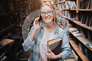 A picture of attractive girl standing with a book in her left hand and talking on the phone. She is looking straight