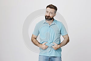 Picture of a bearded handsome man showing his beer belly over white background. Person is bothered because of his extra