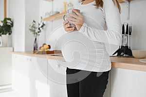 Picture of beautiful casually dressed young brunette female expecting baby, having morning tea in modern stylish kitchen, smiling