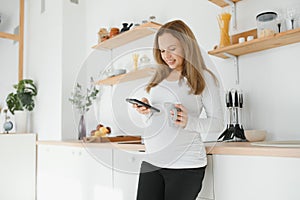 Picture of beautiful casually dressed young brunette female expecting baby, having morning tea in modern stylish kitchen, smiling