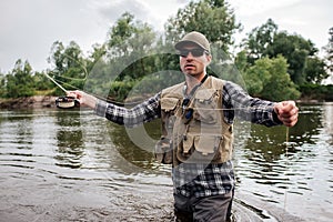 A picture of brutal and calm guy standing in water and looking forward. He has fly rod in one hand and spoon from it in