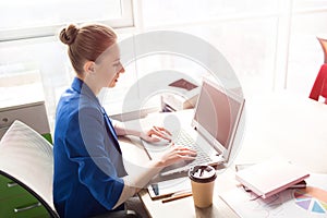 Picture of business woman in blue jacket sitting at the table near bright window and working. She is typing on keybord