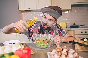 A picture of cook standing at table and holds bottle of sunflower oil. He looks at it and pouring some oil into bowl