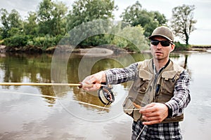 A picture of cool guy stands in water and fishing. He holds fly-fishing with reel under it in one hand and spoon from it
