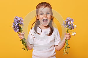 Picture of emotional playful little girl opening mouth and eyes widely, holding flowers in both hands, wearing white t shirt,