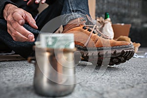 A picture that is focused on boots and a dirty hand of homeless man. He is reaching a cup that stands in front of him
