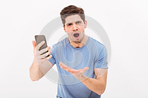 Picture of frustrated unshaved man in casual t-shirt holding smartphone and emotionally reacting on call, isolated over white wall