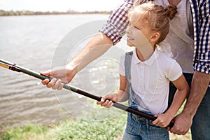 A picture of guy helping his daughter to hold fish-rod in a right way. Girl is holding it with both hands and smiling