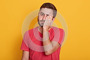Picture of handsome man wearing casual red t shirt, standing with sad expression, covering half of his face with hand, looks tired