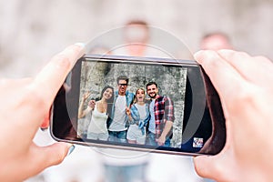 A picture of human`s hands holding phone. This human is taking picture of four happy people. They are standing on grey