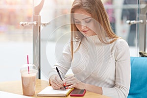 Picture of inspired attractive model writing at her notebook, spending time alone in cafe, having smartphone and cup of cocktail