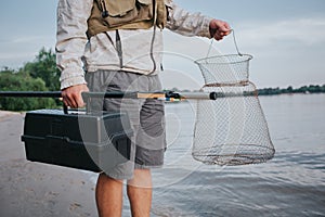 A picture of man holding fishing net and plastic black box in hands. Also he has fly rold in the right one. Guy is