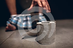 Picture of man`s hand reaching to paper roll after defecation. Guy sit on pot in rest room with pants on his feet. Alone