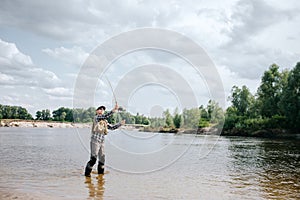 A picture of man standing in water and waving with fly rod. He holds with both hands. Guy is looking at fly rod. He