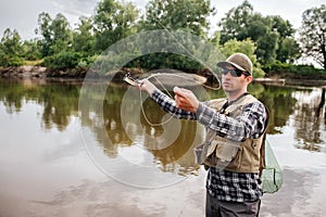 A picture of man stands in water and waving with fly rod. He is going to fish. Guy has fishing net on the back. He looks