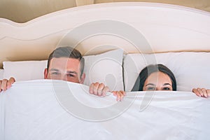A picture of man and woman hiding faces under white blanket. They are lying together in one bed. Peple are looking up